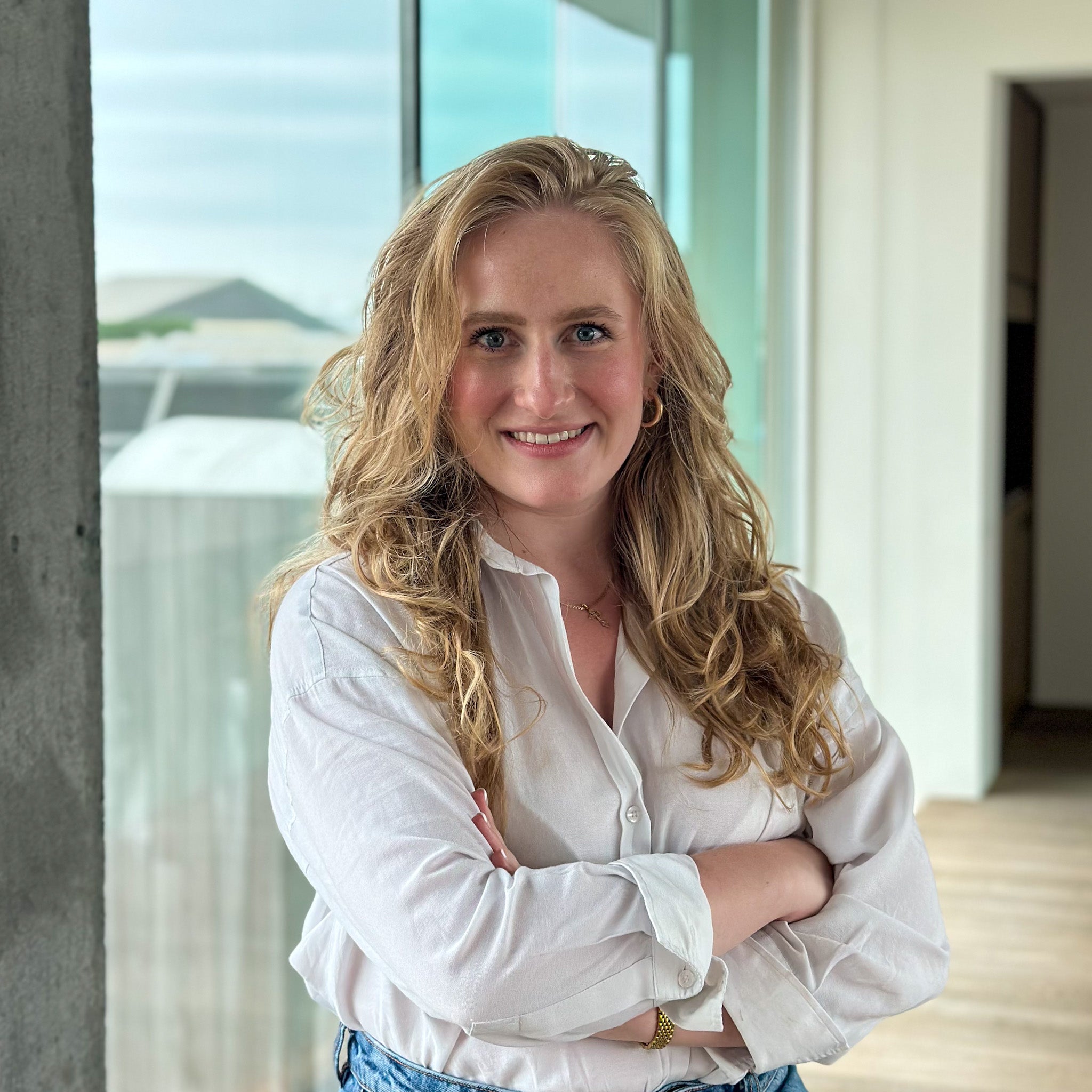 Smiling blonde woman in a white button-down shirt and jeans, arms crossed, in a modern office.