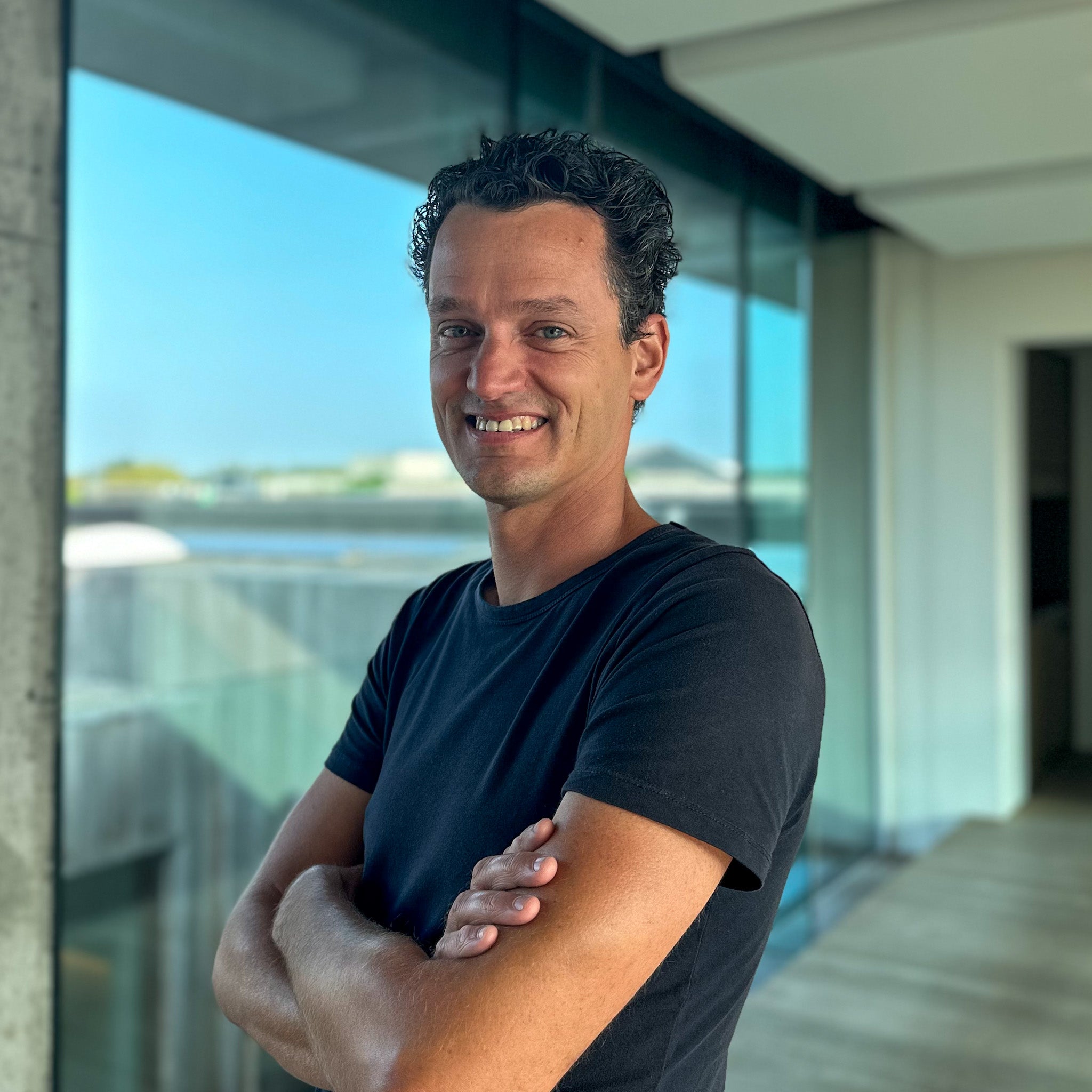 Smiling man with dark curly hair and a black t-shirt, arms crossed, in a modern building with large windows.