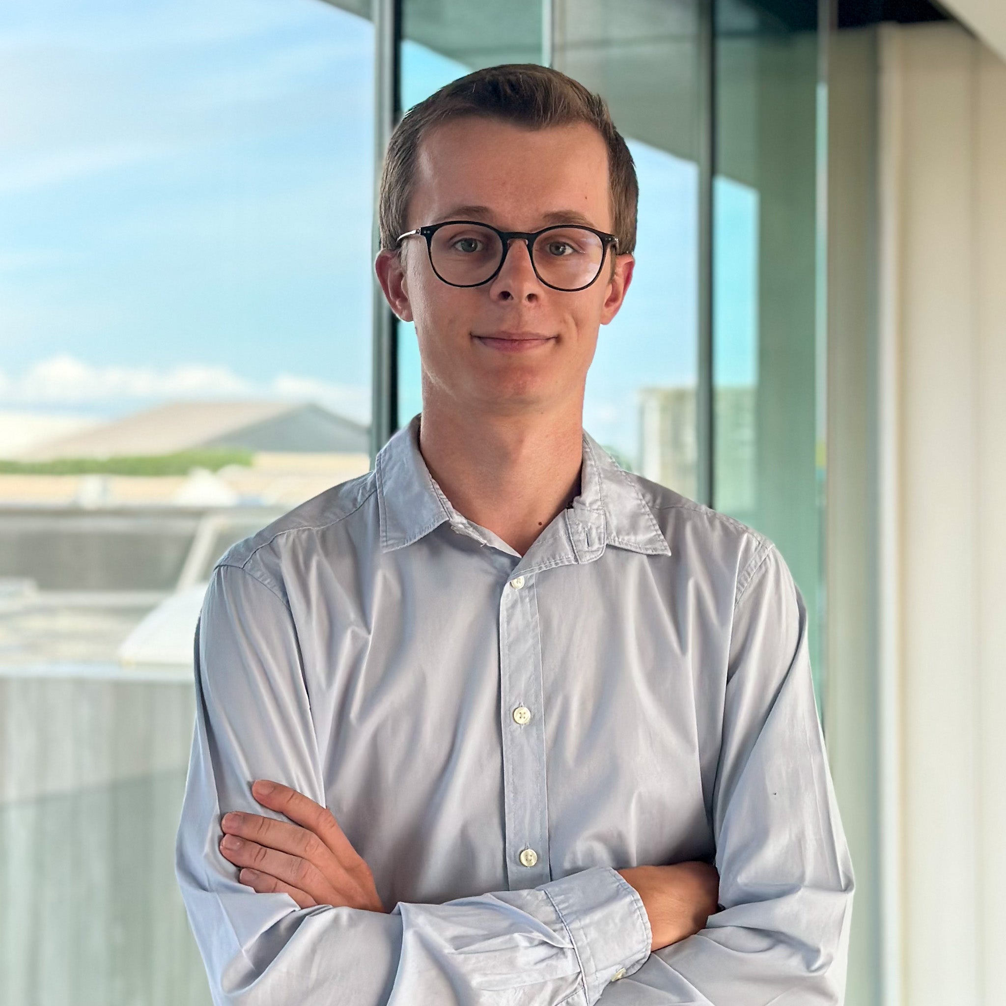 Young man with glasses and a light blue shirt, arms crossed, standing in front of a window.