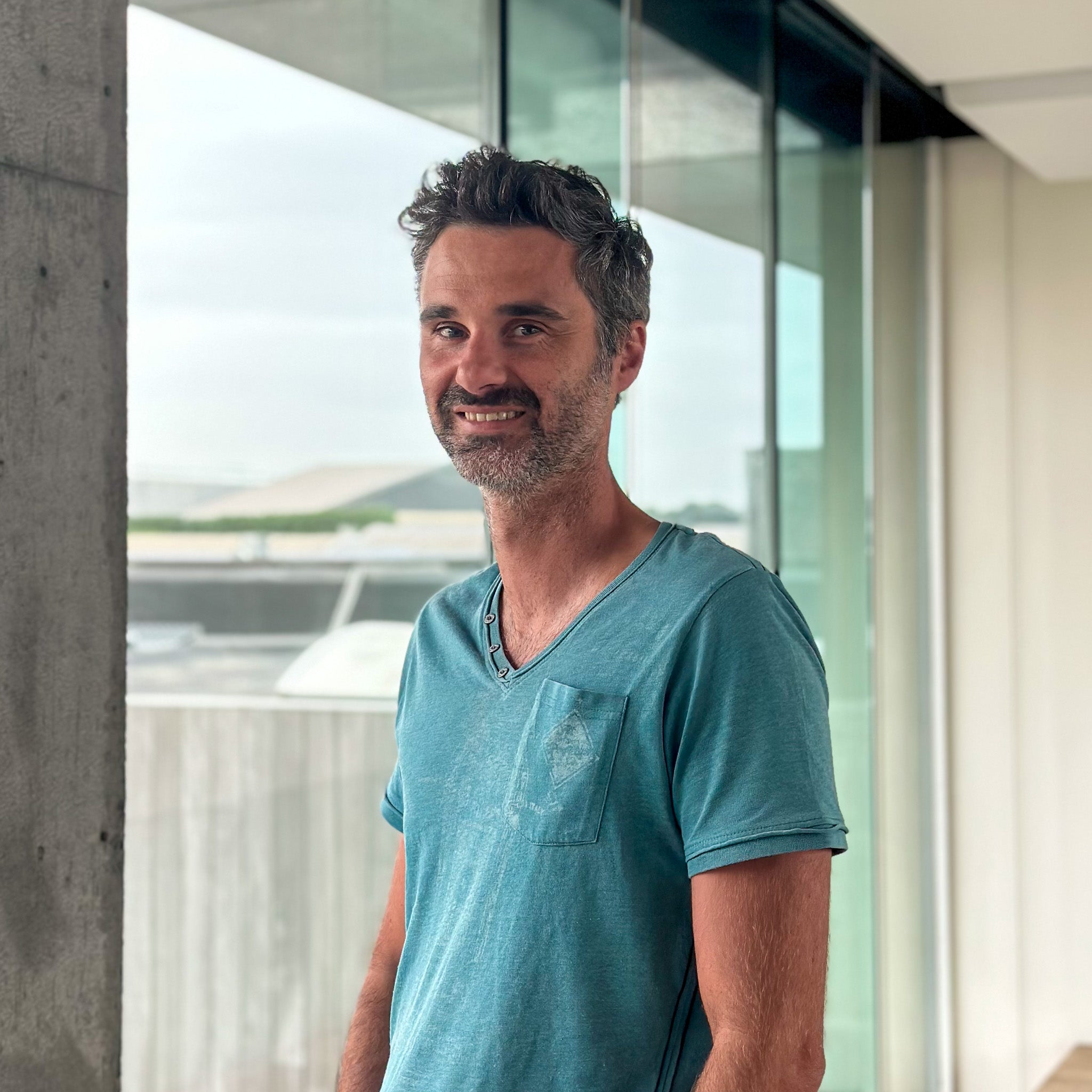 Smiling man with dark, graying hair and a beard, wearing a teal v-neck shirt, standing by a large window.