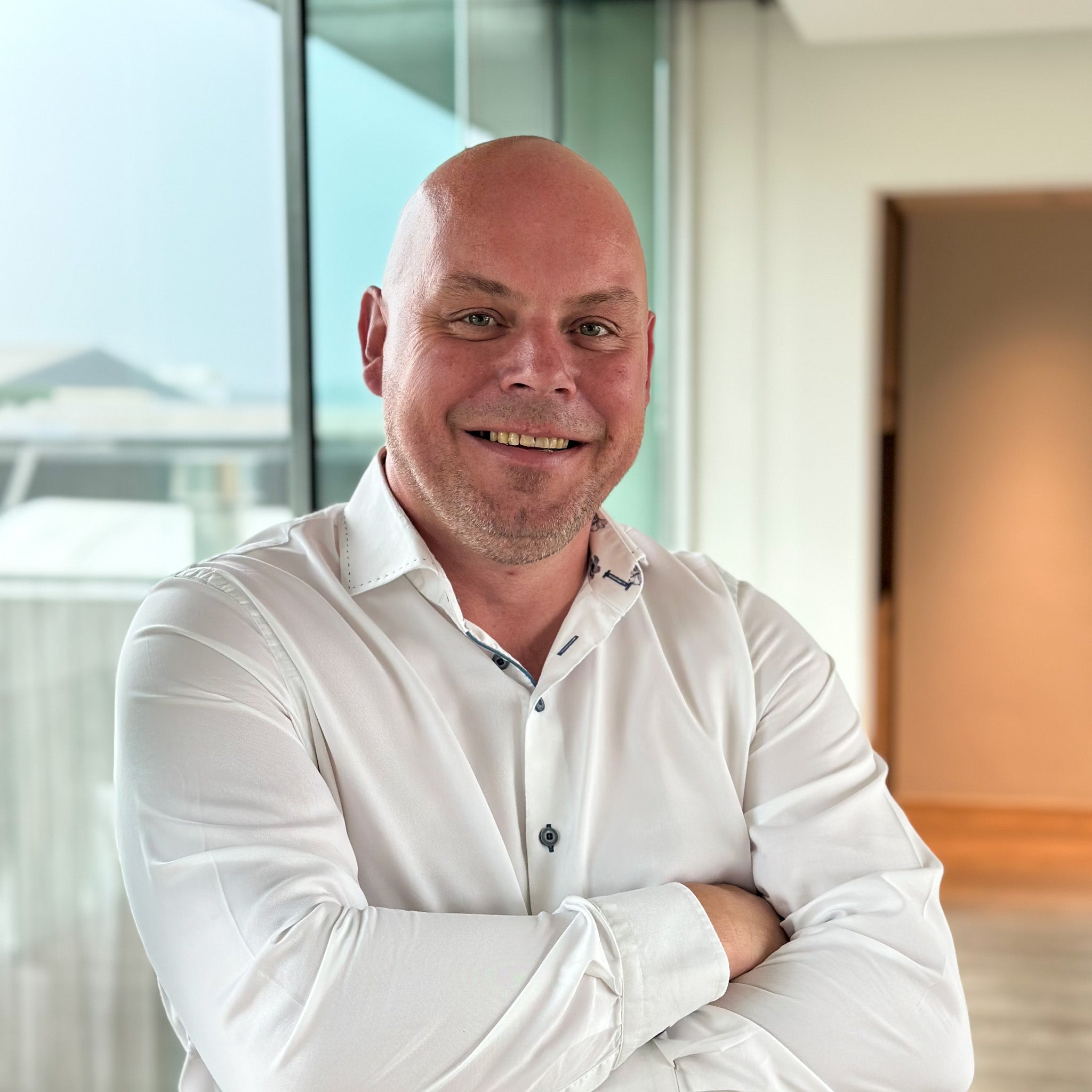 Smiling bald man in a white collared shirt with arms crossed, standing indoors by a large window.