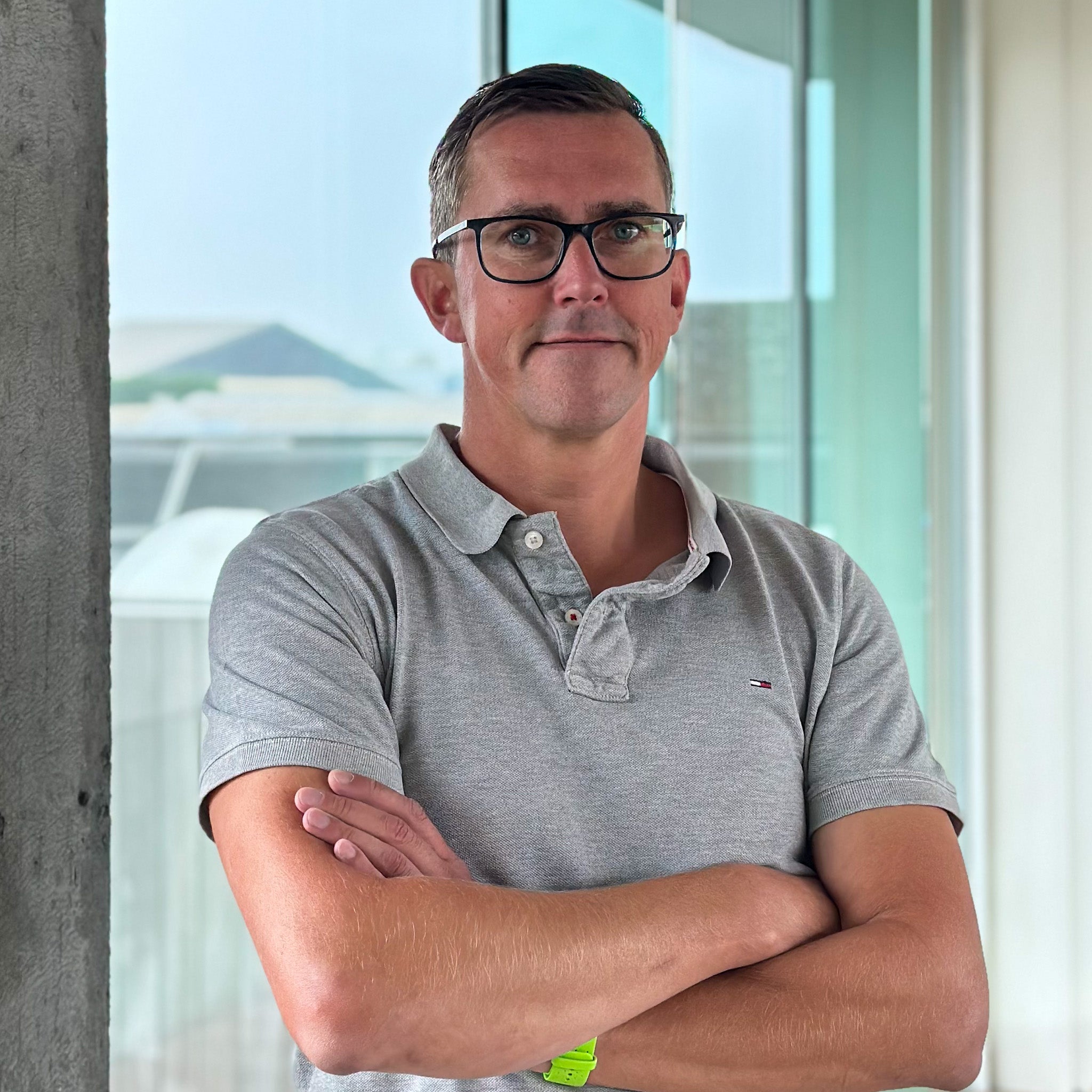 Man in gray polo shirt and glasses, arms crossed, standing in front of a window.