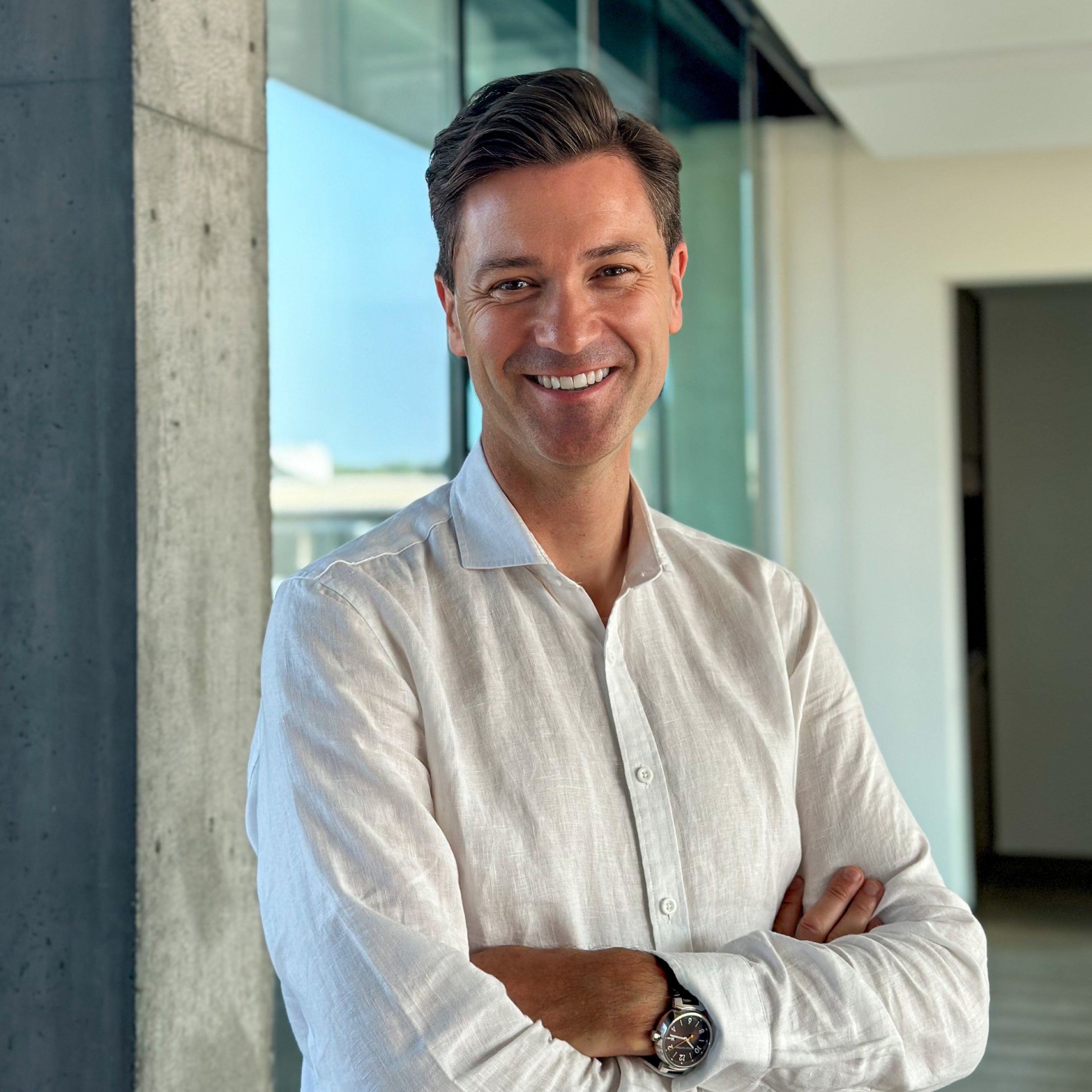 Smiling man in a white linen shirt with arms crossed, standing in a modern office with glass walls.