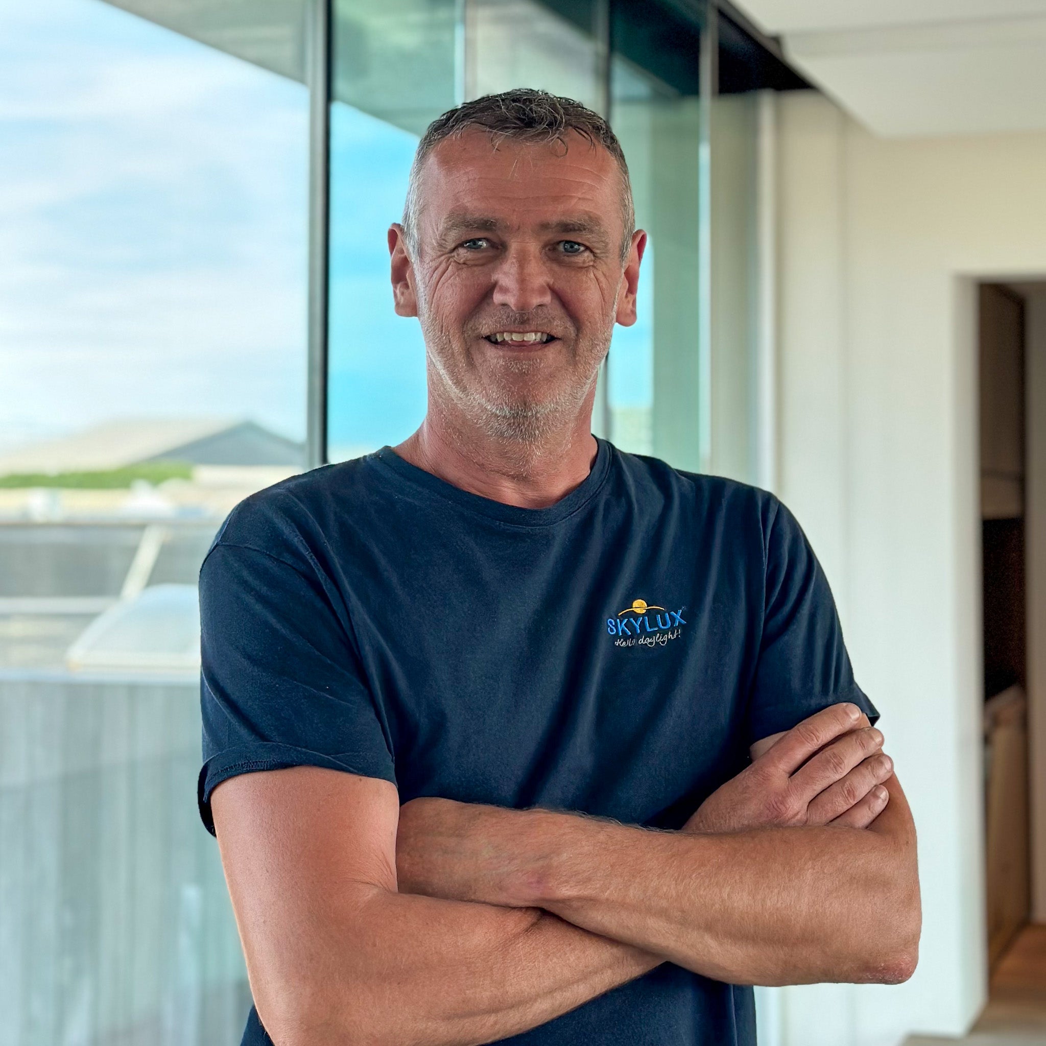 Smiling man with gray hair in a navy "Skylux" t-shirt, arms crossed, against a bright window.
