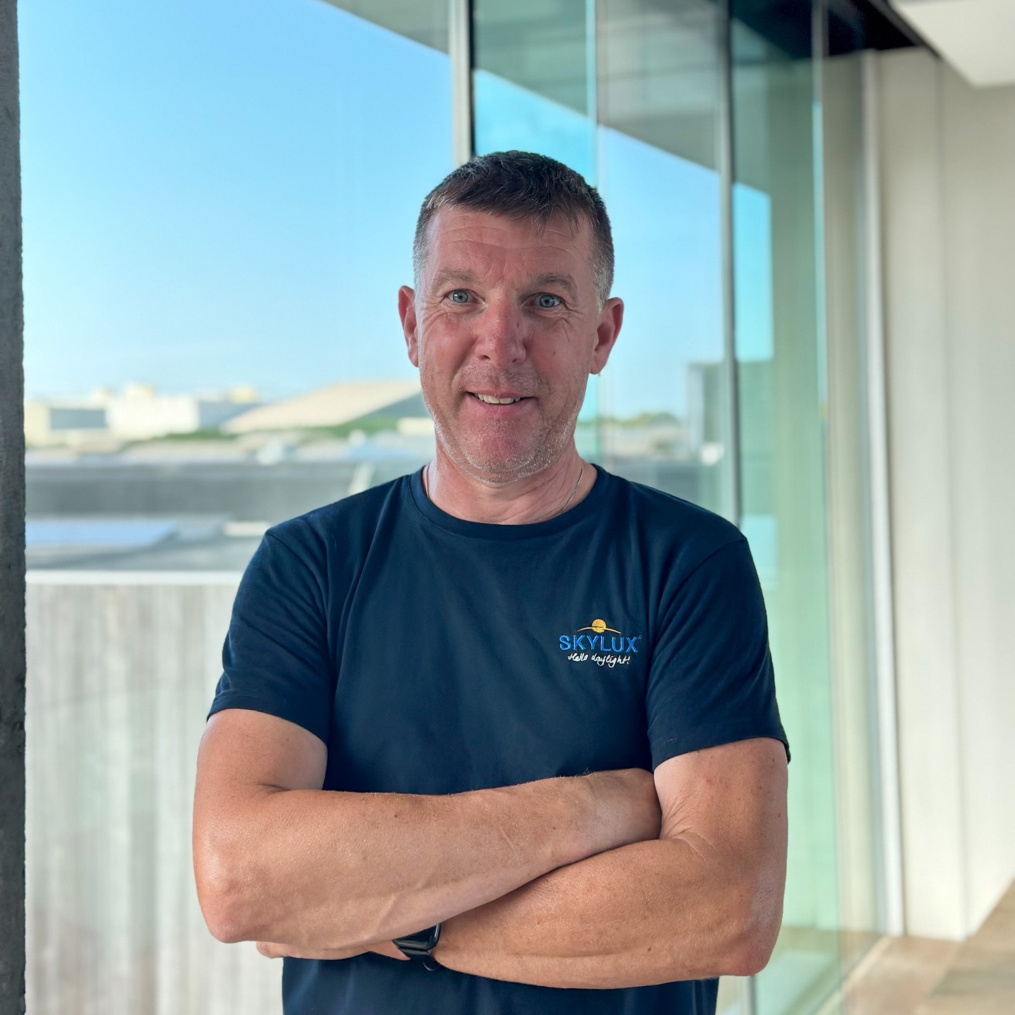 Man in a dark blue t-shirt with "SKYLUX" logo, arms crossed, smiling in front of a window.