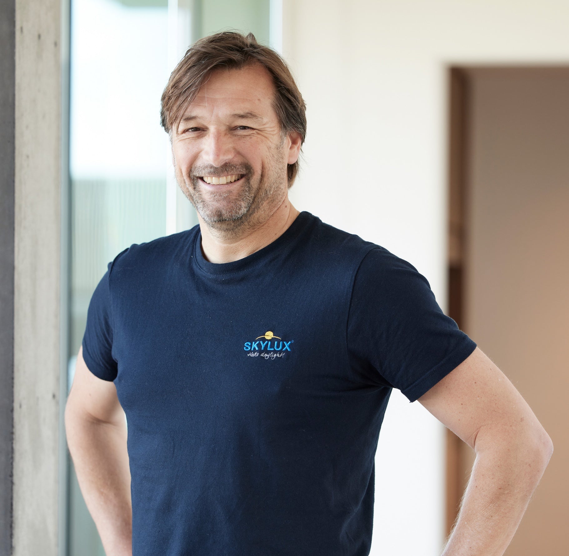 Smiling man in a navy blue Skylux t-shirt, standing indoors with hands on hips.