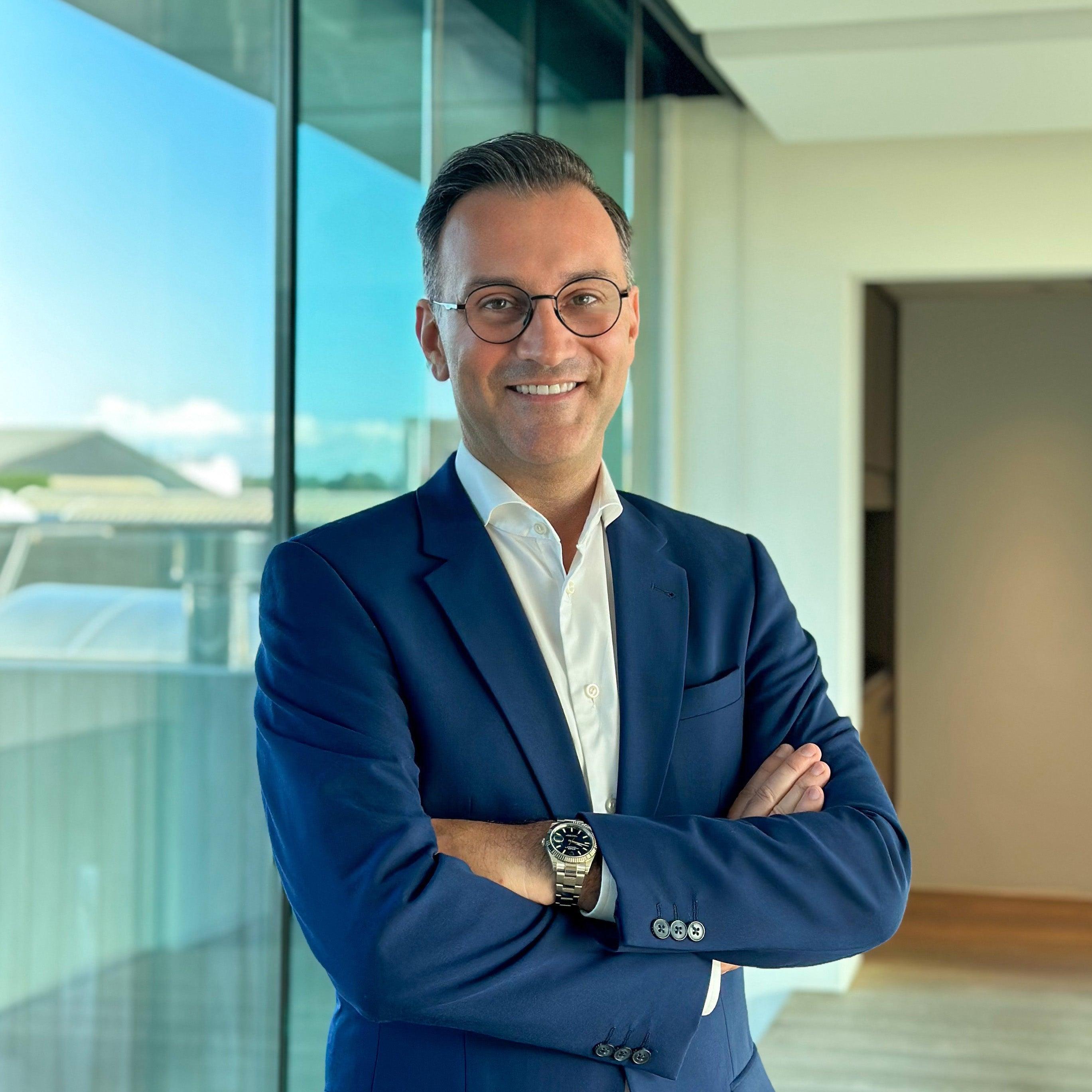 Smiling man in a blue suit and glasses, arms crossed, standing in a modern office with large windows.