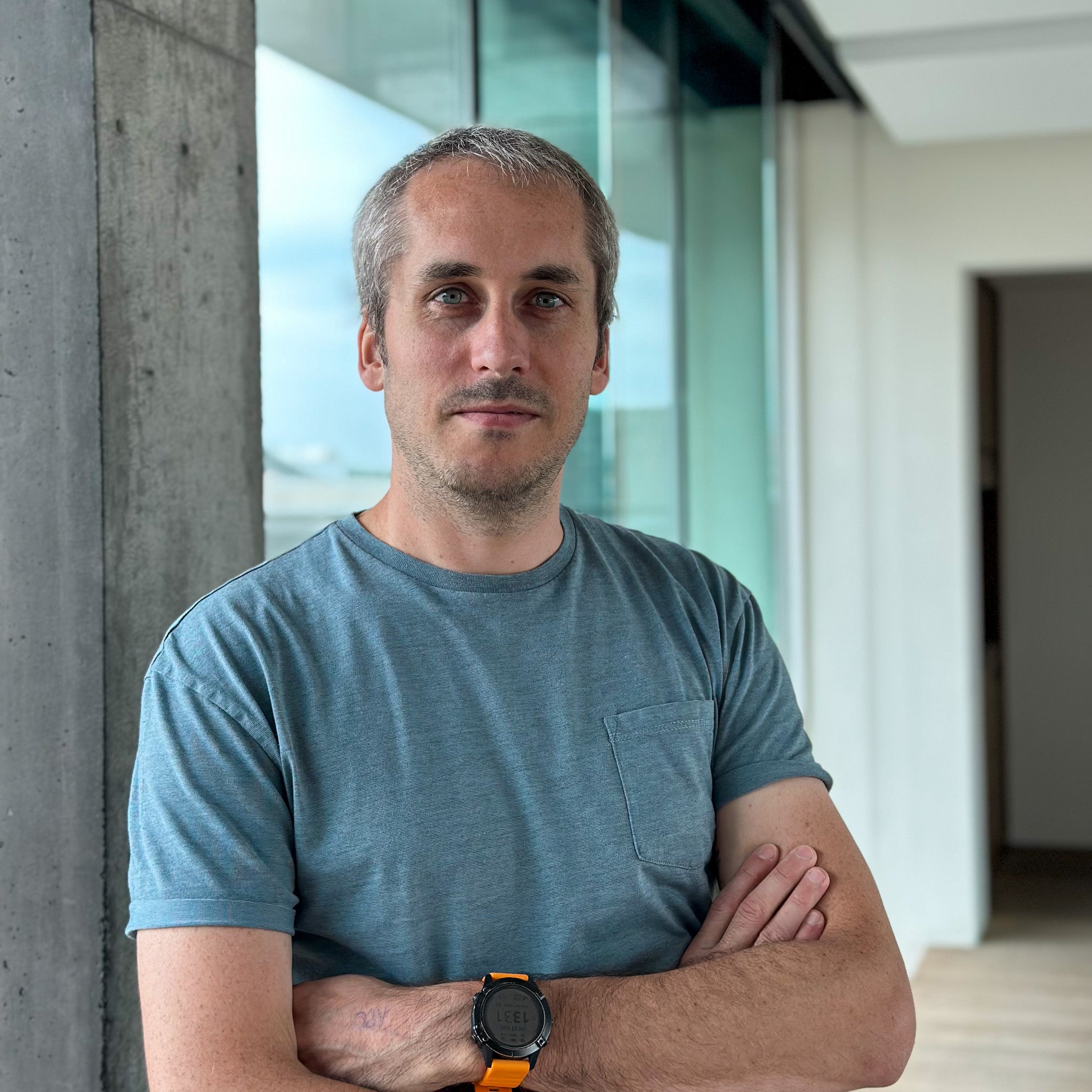 Man with graying hair in a blue t-shirt and orange smartwatch, arms crossed, against a modern concrete and glass background.