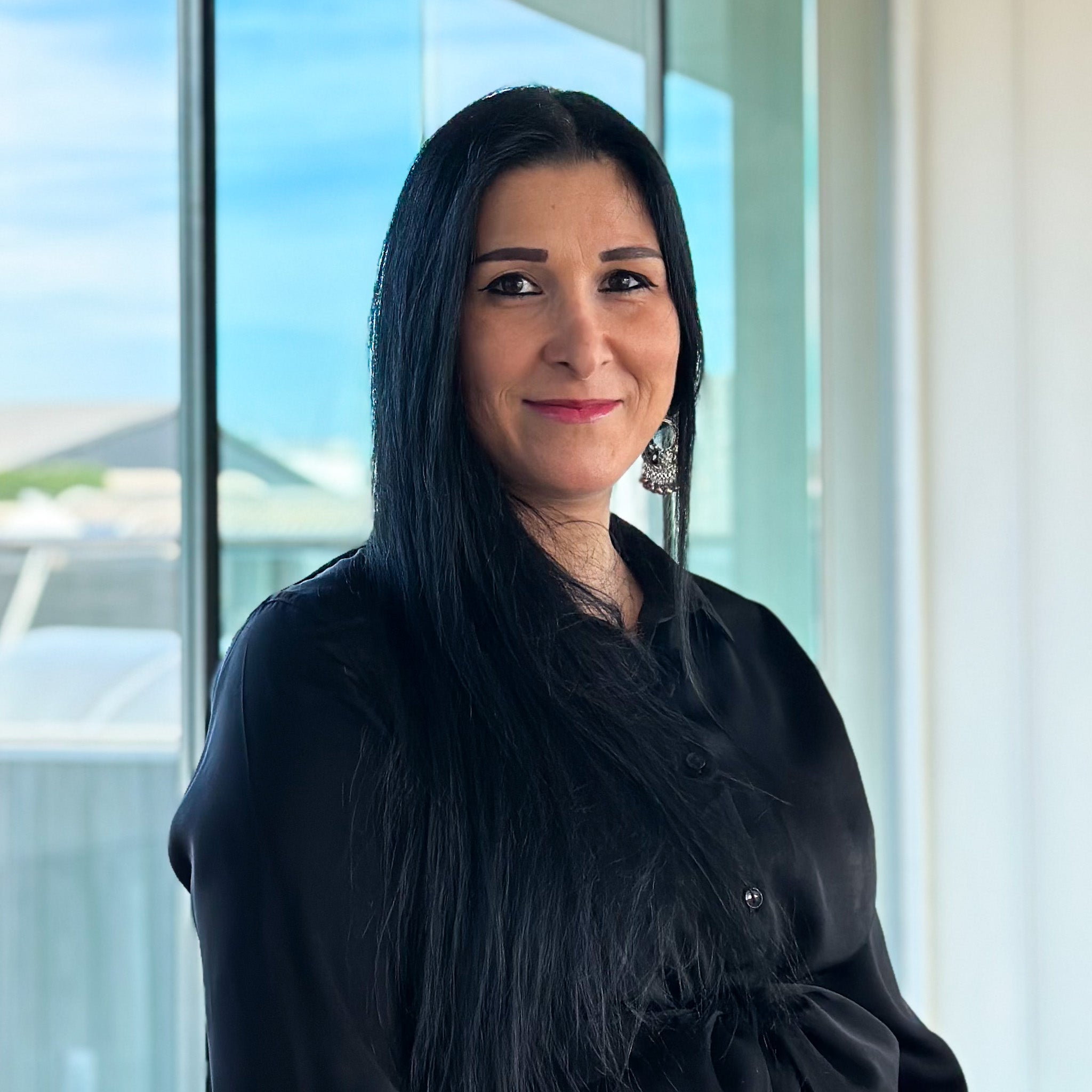 Jessica Mezzina, a woman with long black hair, wearing a black shirt and ornate earrings, smiles against a bright window.
