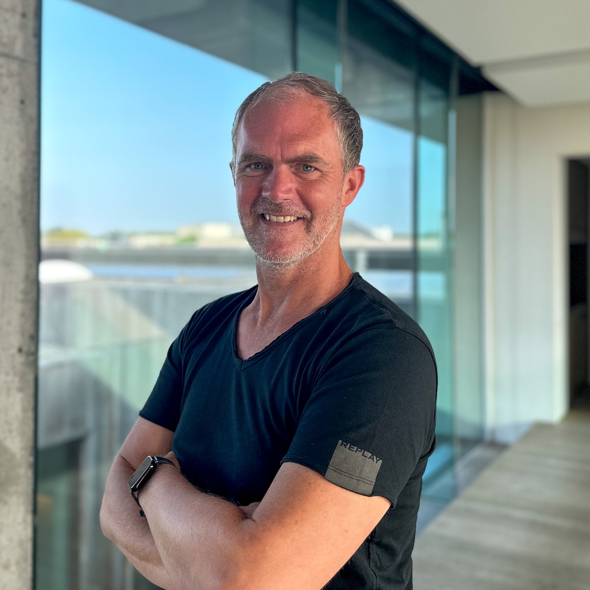 Smiling man with graying hair, black t-shirt, and smartwatch, arms crossed, in a modern building.
