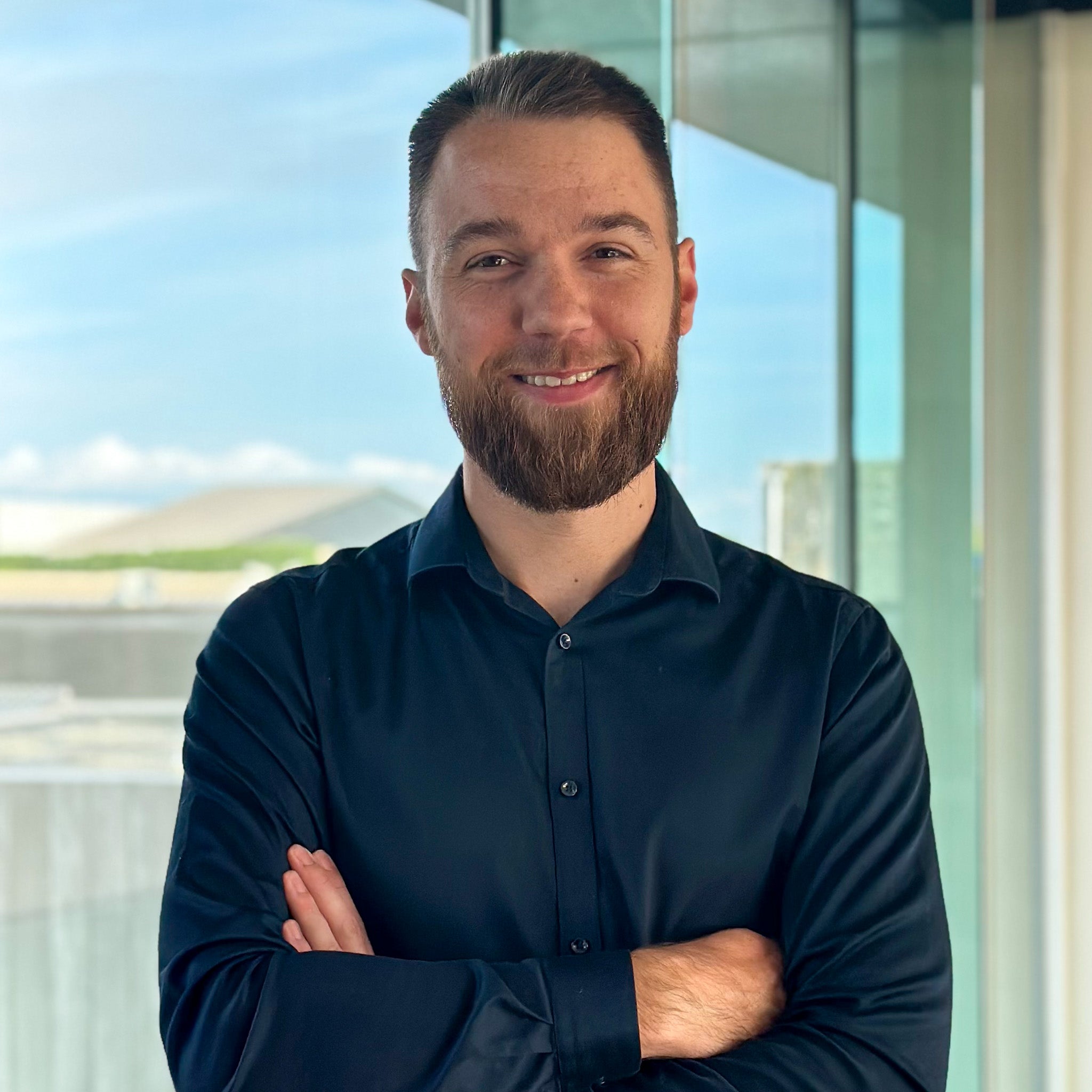 Smiling man with a beard in a dark blue shirt, arms crossed, standing by a window.