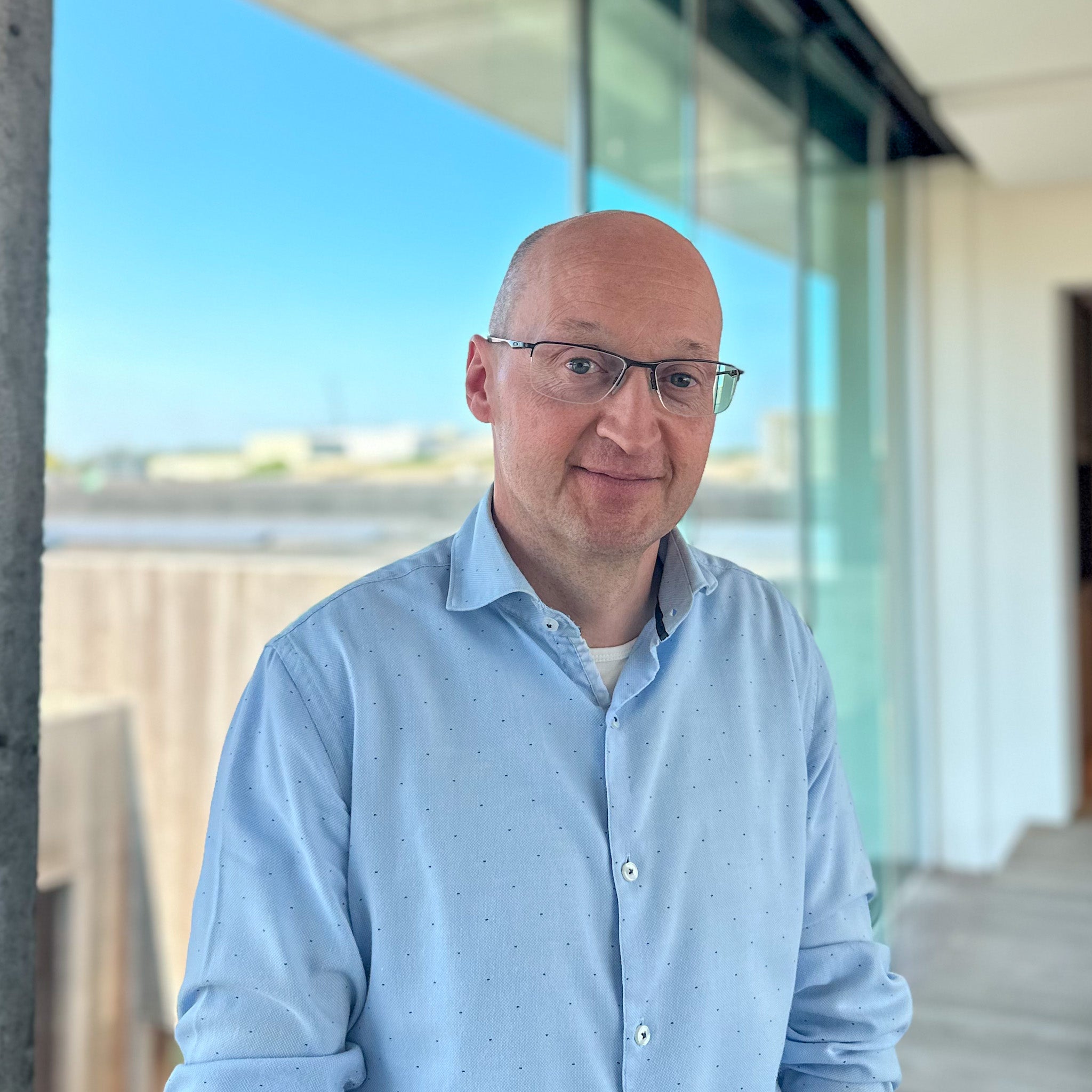 Jurgen Voet, a bald man in glasses and a light blue shirt, stands outdoors with a modern building and blue sky behind him.