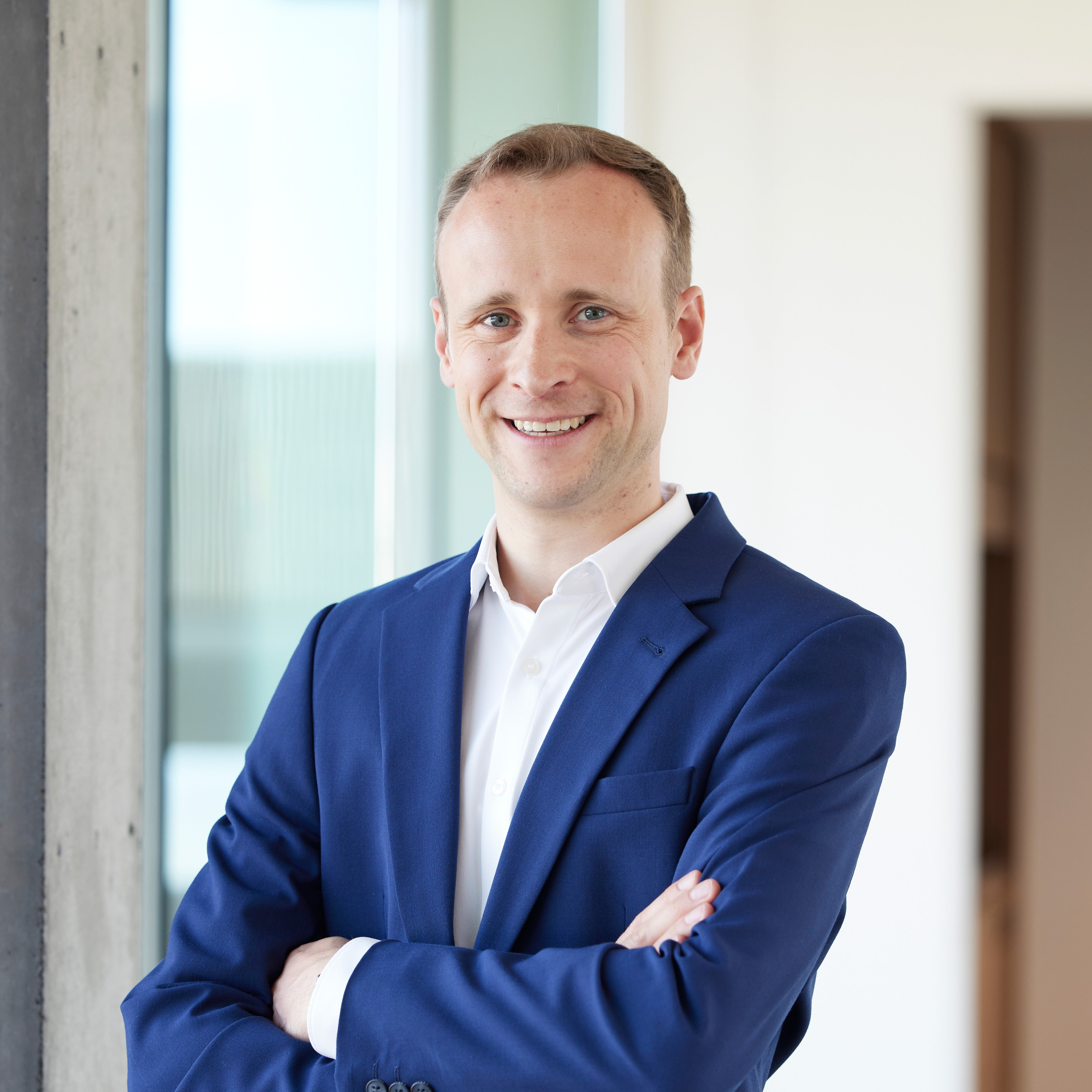 Smiling man in a sharp blue suit and white shirt, arms crossed, in a modern office setting.