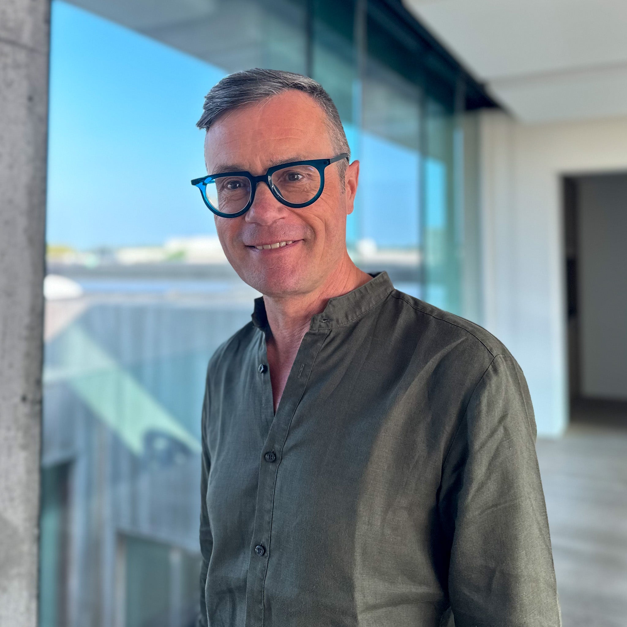 Smiling man with gray hair and blue glasses wearing a dark olive green shirt, standing indoors by a window.
