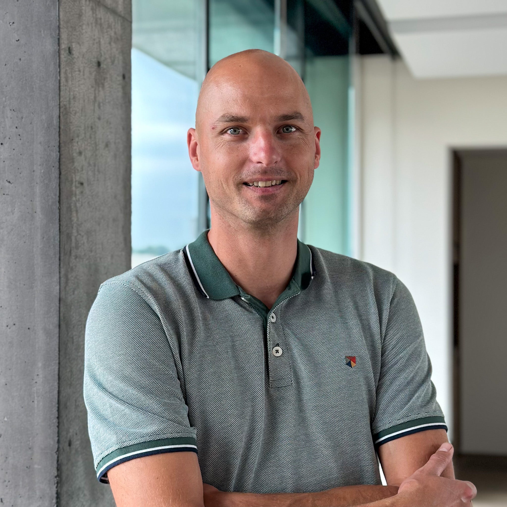Maarten Deheegher, a bald man in a green polo shirt, smiles with arms crossed in a modern office.