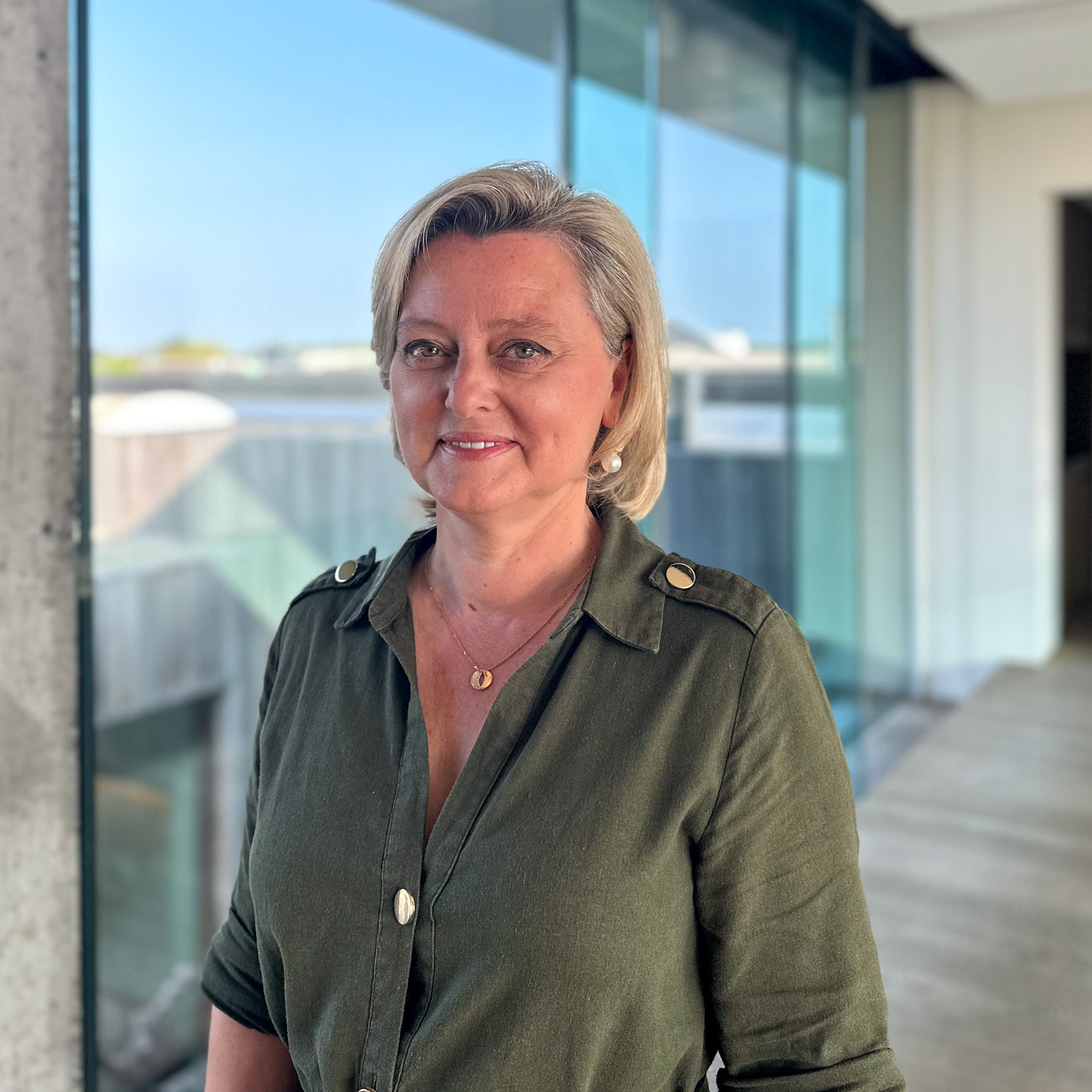 Blonde woman in an olive green shirt with gold buttons and necklace, smiling in front of a glass window.