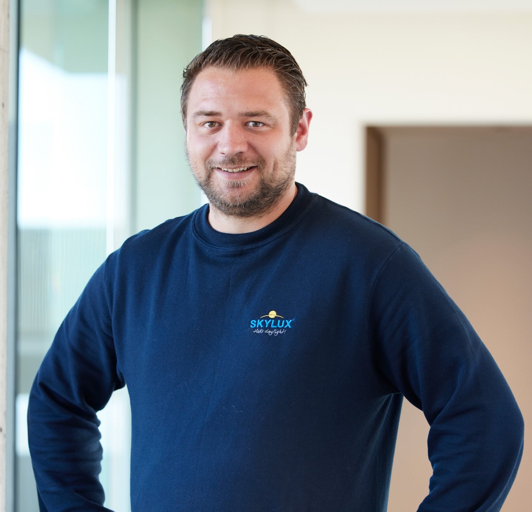 Mathieu Bruyneel, a smiling man in a navy blue Skylux sweatshirt, stands indoors.