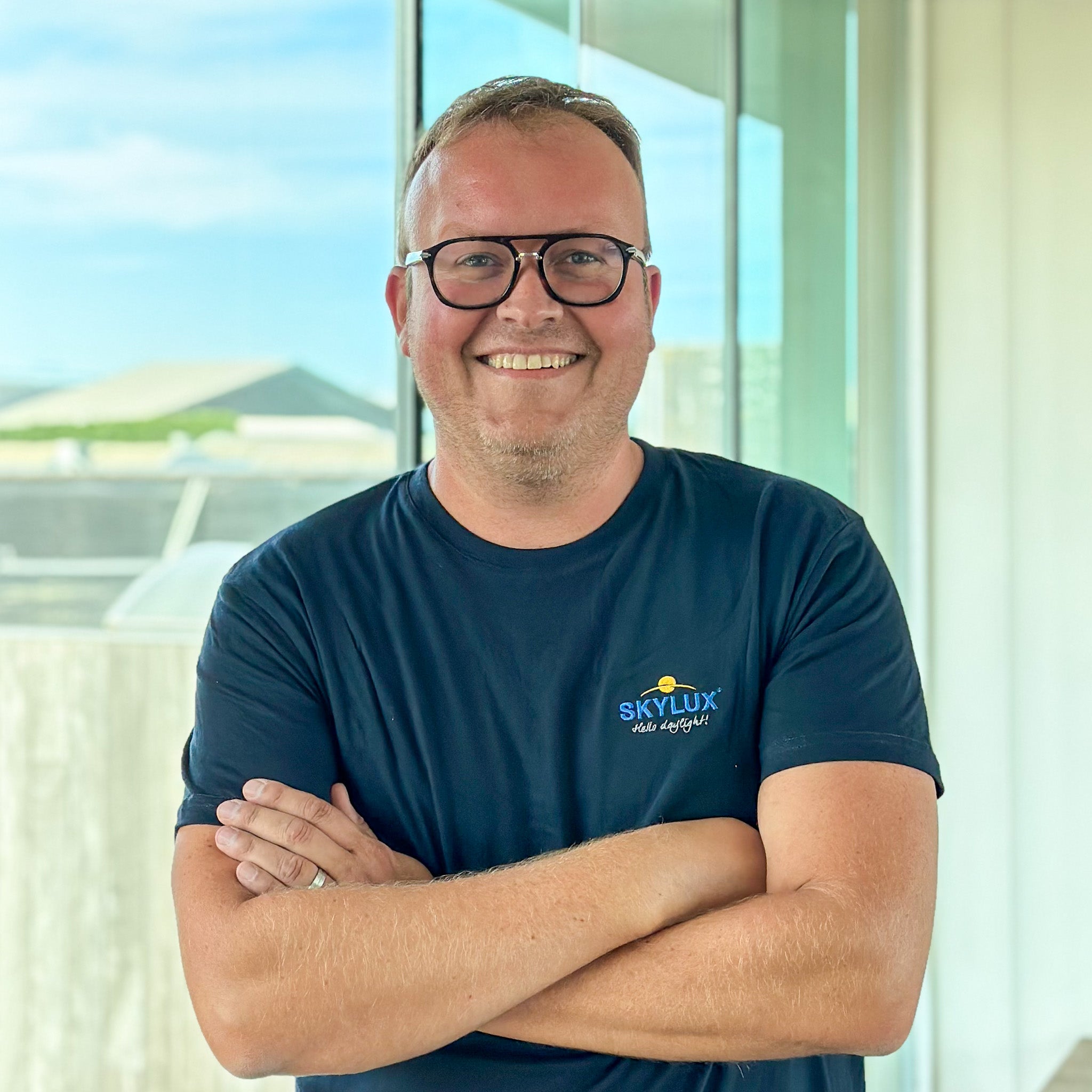 Smiling man in black-rimmed glasses and a navy "SKYLUX" t-shirt, arms crossed, against a bright window.
