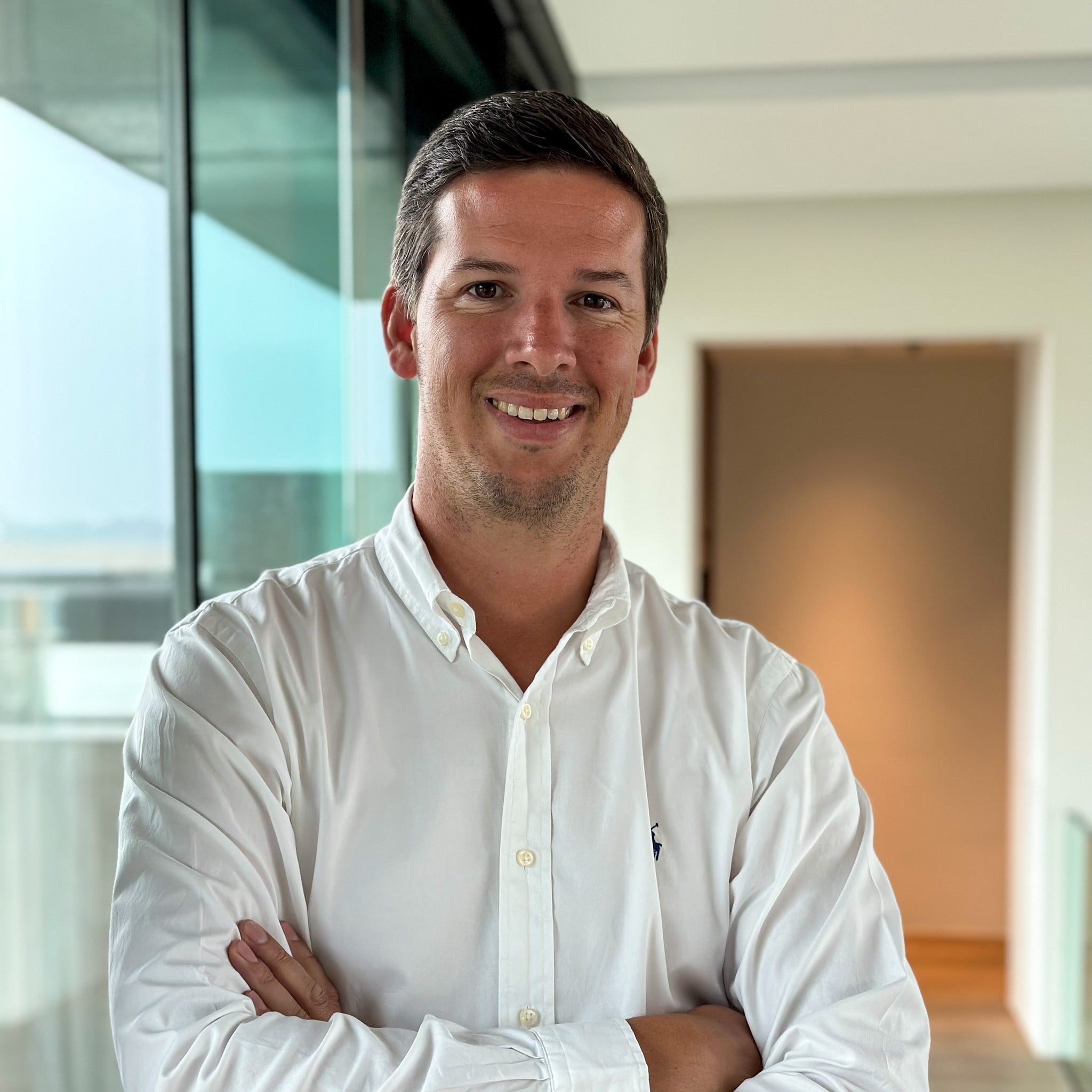 Smiling man in a white button-down shirt with arms crossed, standing in a modern office with glass walls.