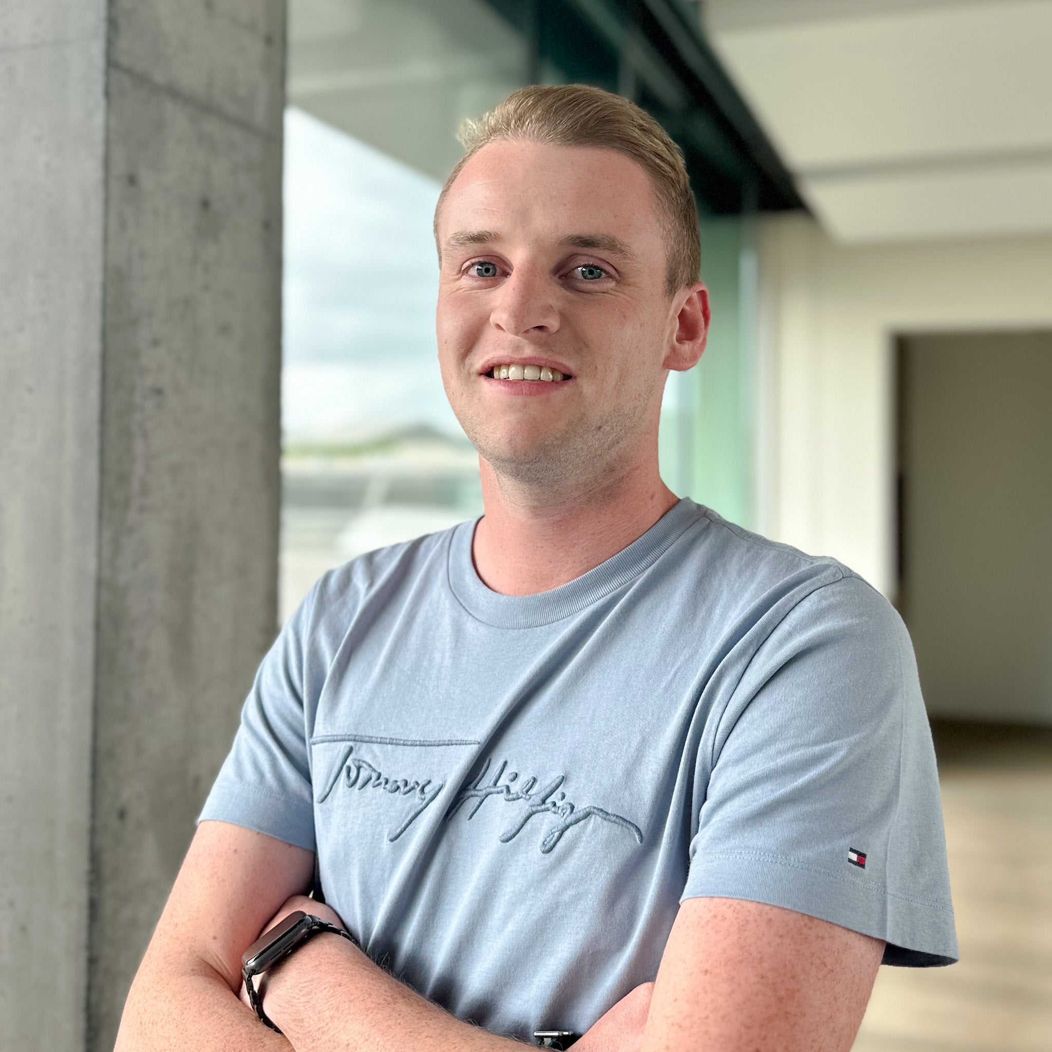 Young man with blonde hair and blue eyes, wearing a light blue Tommy Hilfiger t-shirt, smiling with arms crossed.
