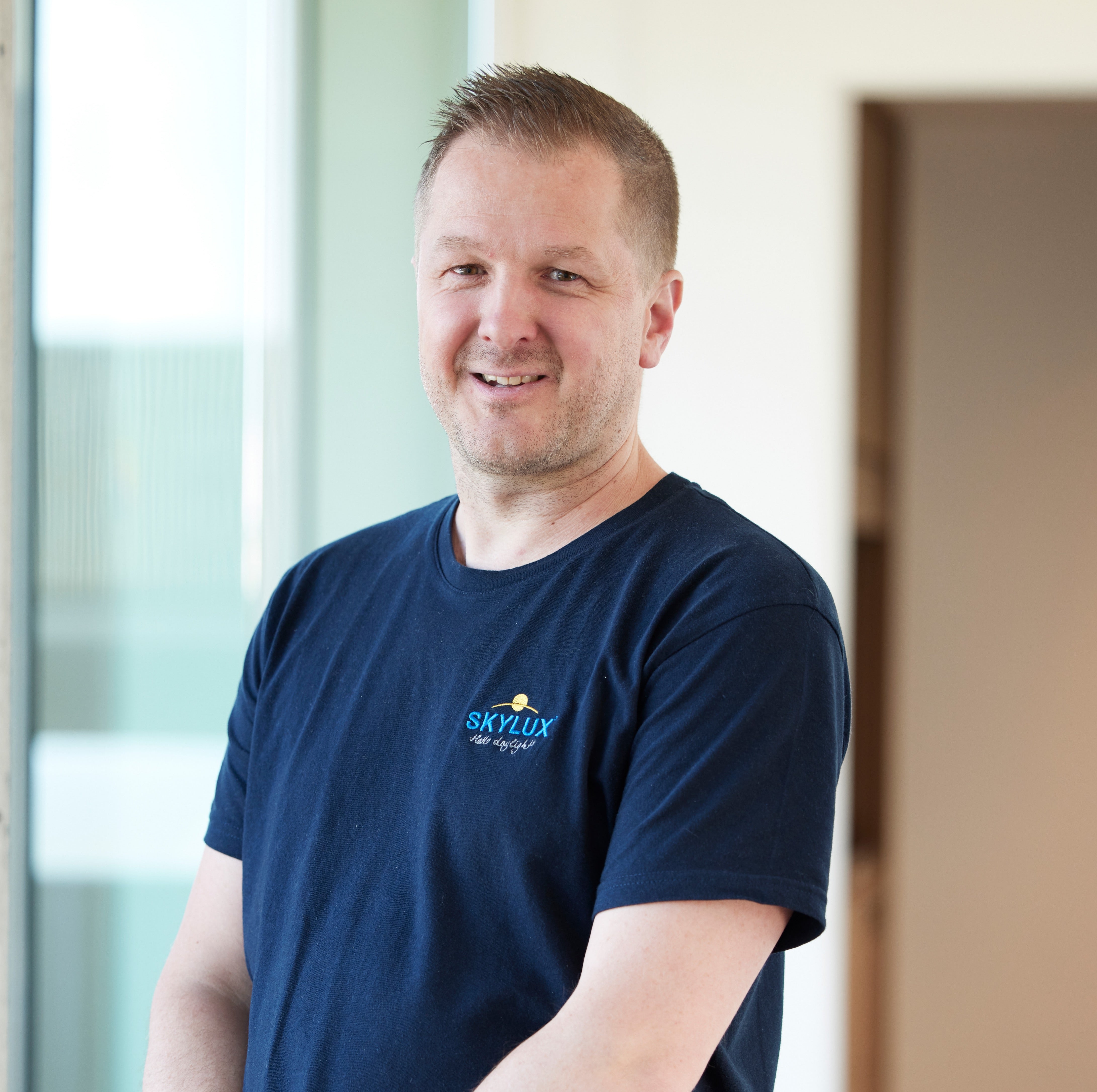 Man in a navy blue Skylux t-shirt smiling at the camera, standing indoors with natural light.