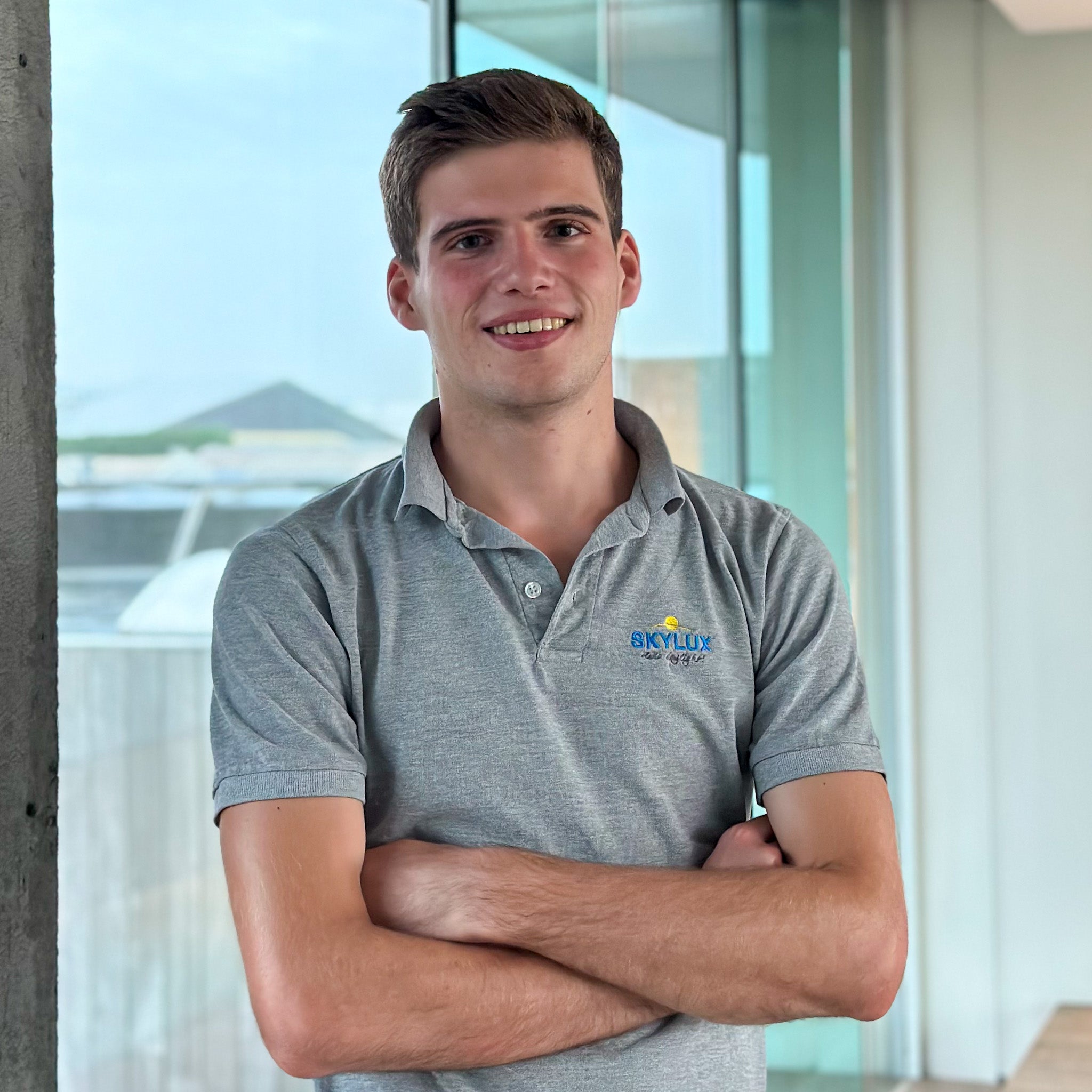 Smiling young man in a grey polo shirt with a "SKYLUX" logo, arms crossed, standing by a window.