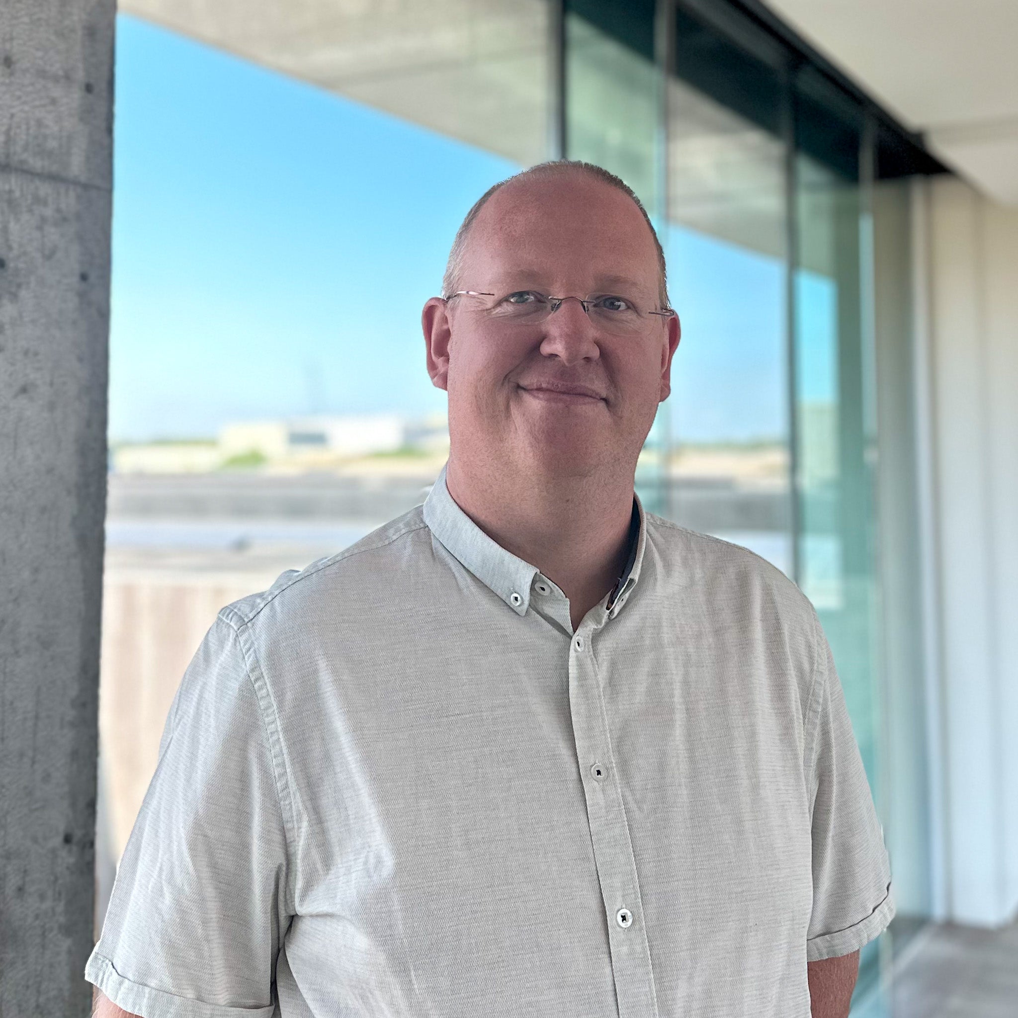 Tom Braeckevelt, a man in a light grey shirt and glasses, smiles in front of a large window.