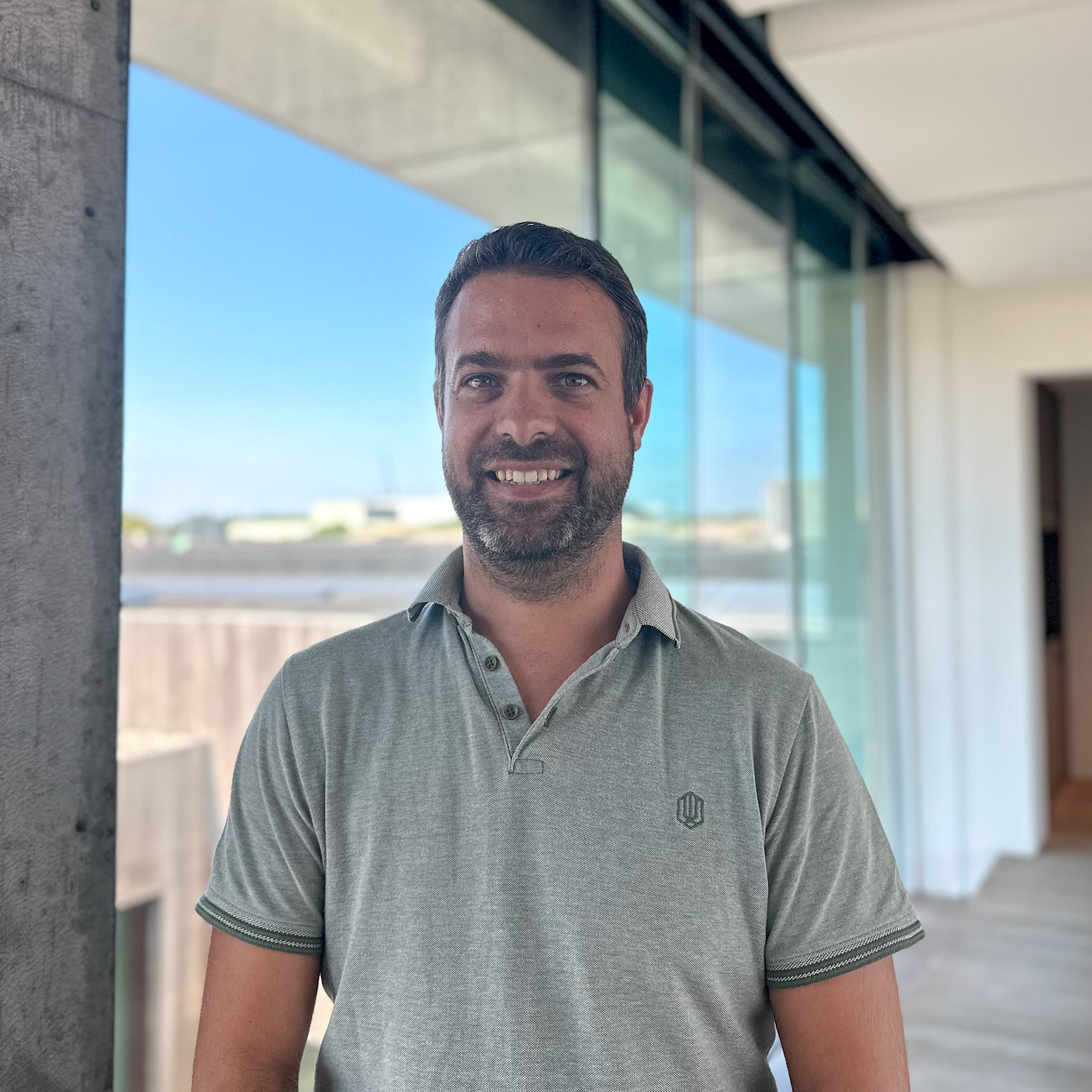 Smiling man with dark hair and beard in a grey polo shirt, standing indoors by a large window.