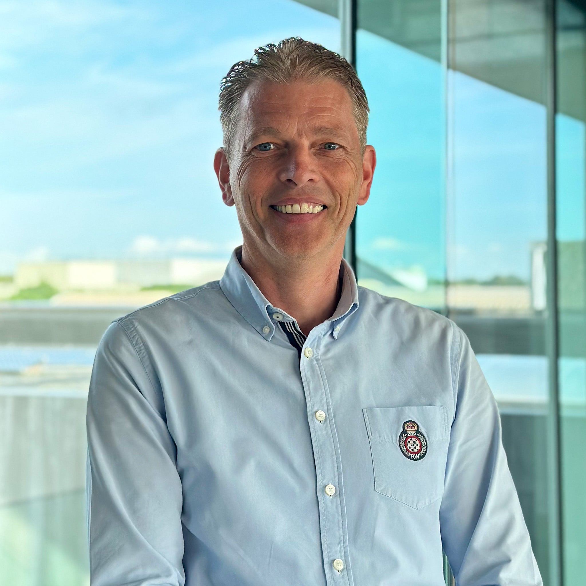 Tony Destrooper, a smiling man in a light blue button-down shirt with a crest logo, stands by a window.
