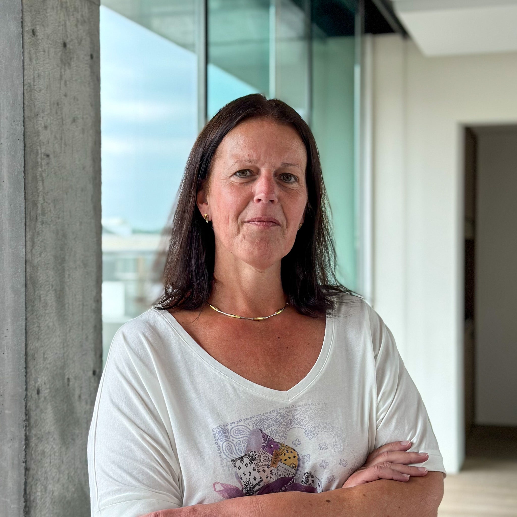 Veerle Verslycke, a woman with dark hair, wearing a white t-shirt and gold necklace, arms crossed.
