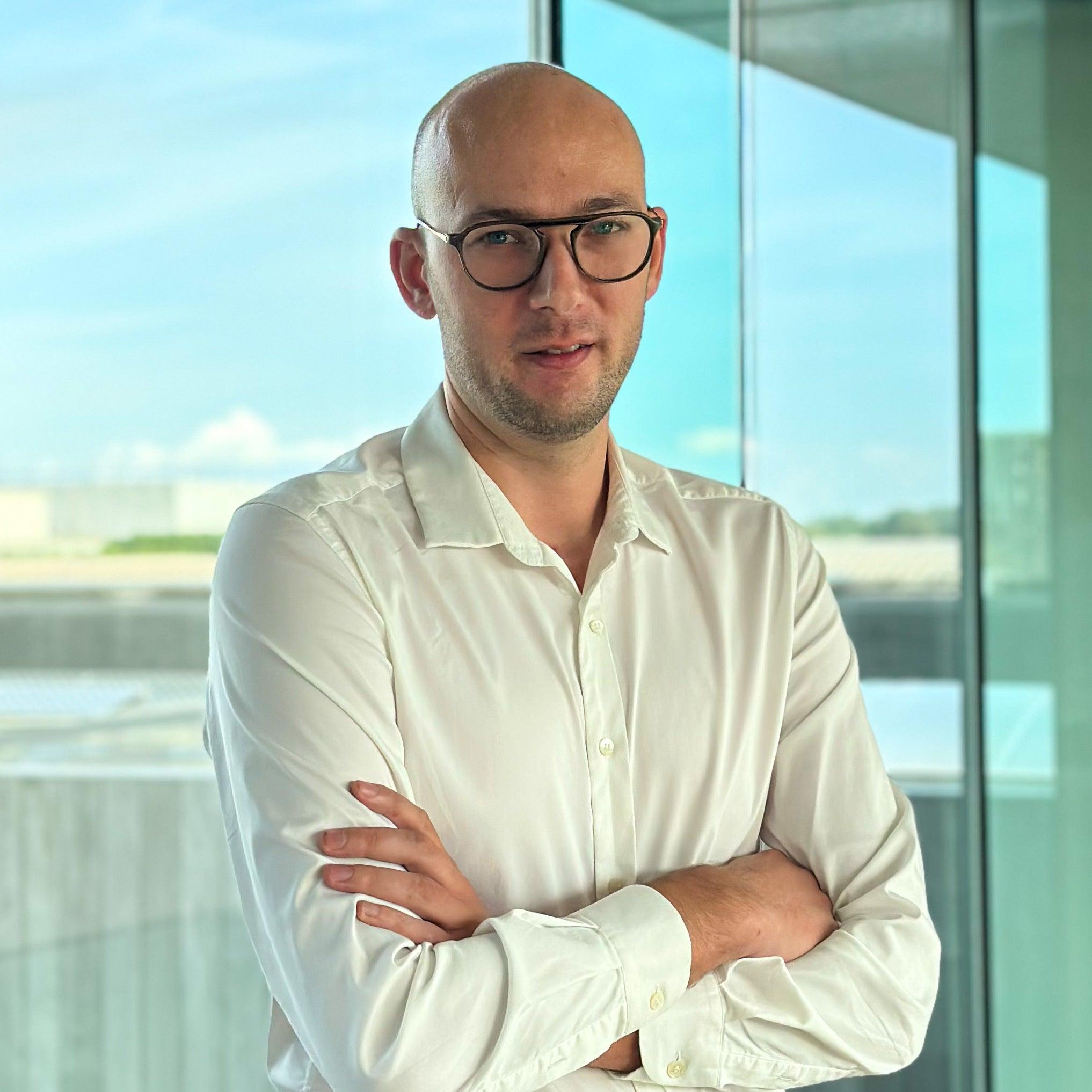 Bald man in glasses and white shirt, arms crossed, standing before a modern window.
