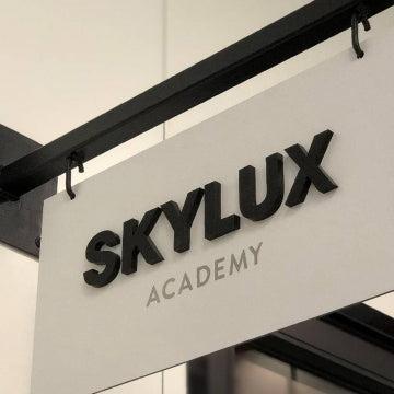White hanging sign with black "SKYLUX ACADEMY" text, mounted on a dark metal bracket.